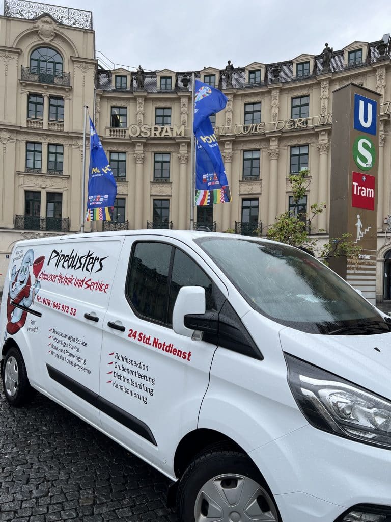 Ein Pipebusters Servicefahrzeug geparkt in München vor einem historischen Gebäude mit blauen Fahnen und U-Bahn-Symbol.